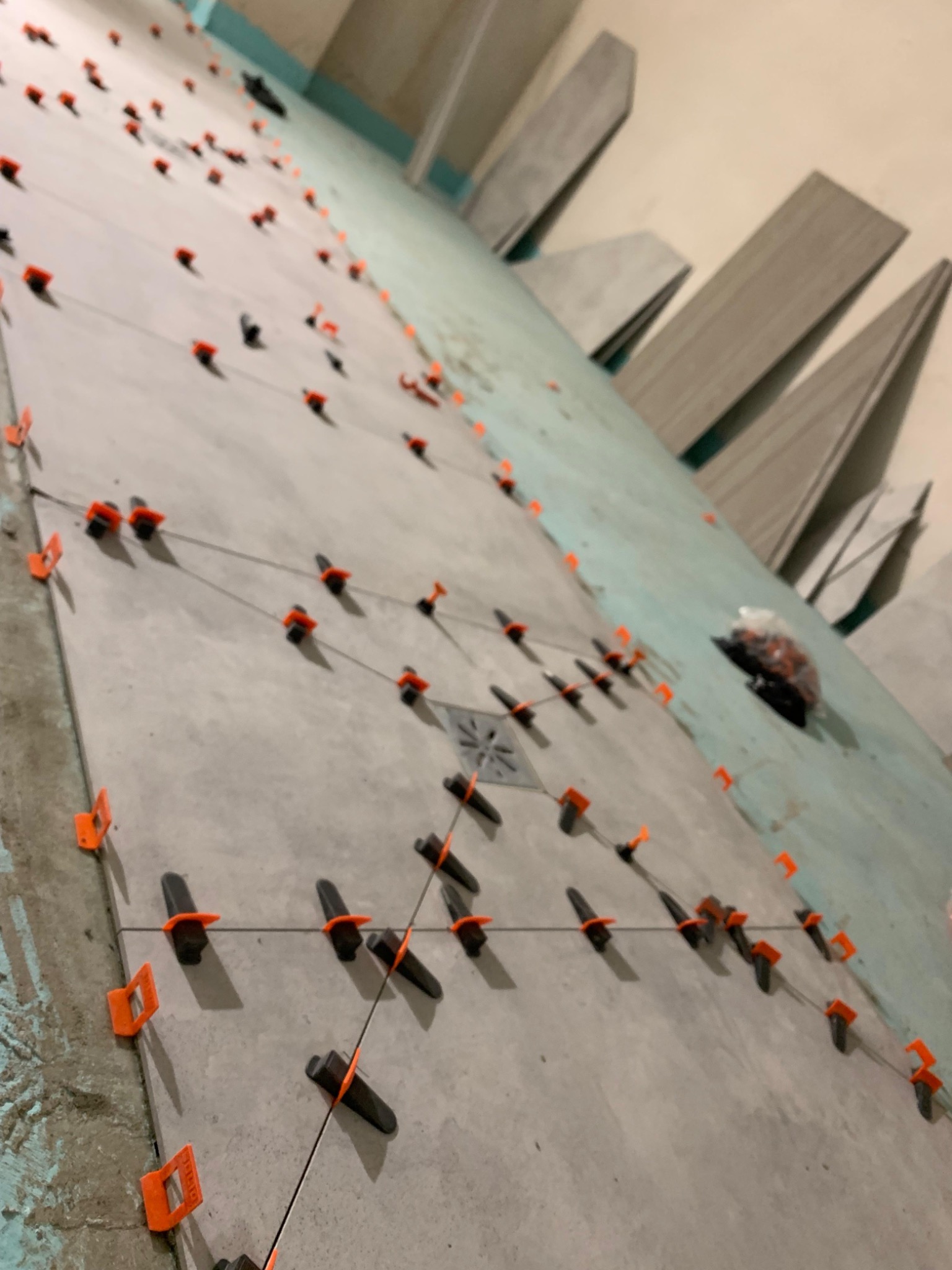 Szare płytki podłogowe z systemem poziomowania, widoczny odpływ liniowy. Płytki ułożone do ściany, na której opierają się pionowo ustawione płytki.