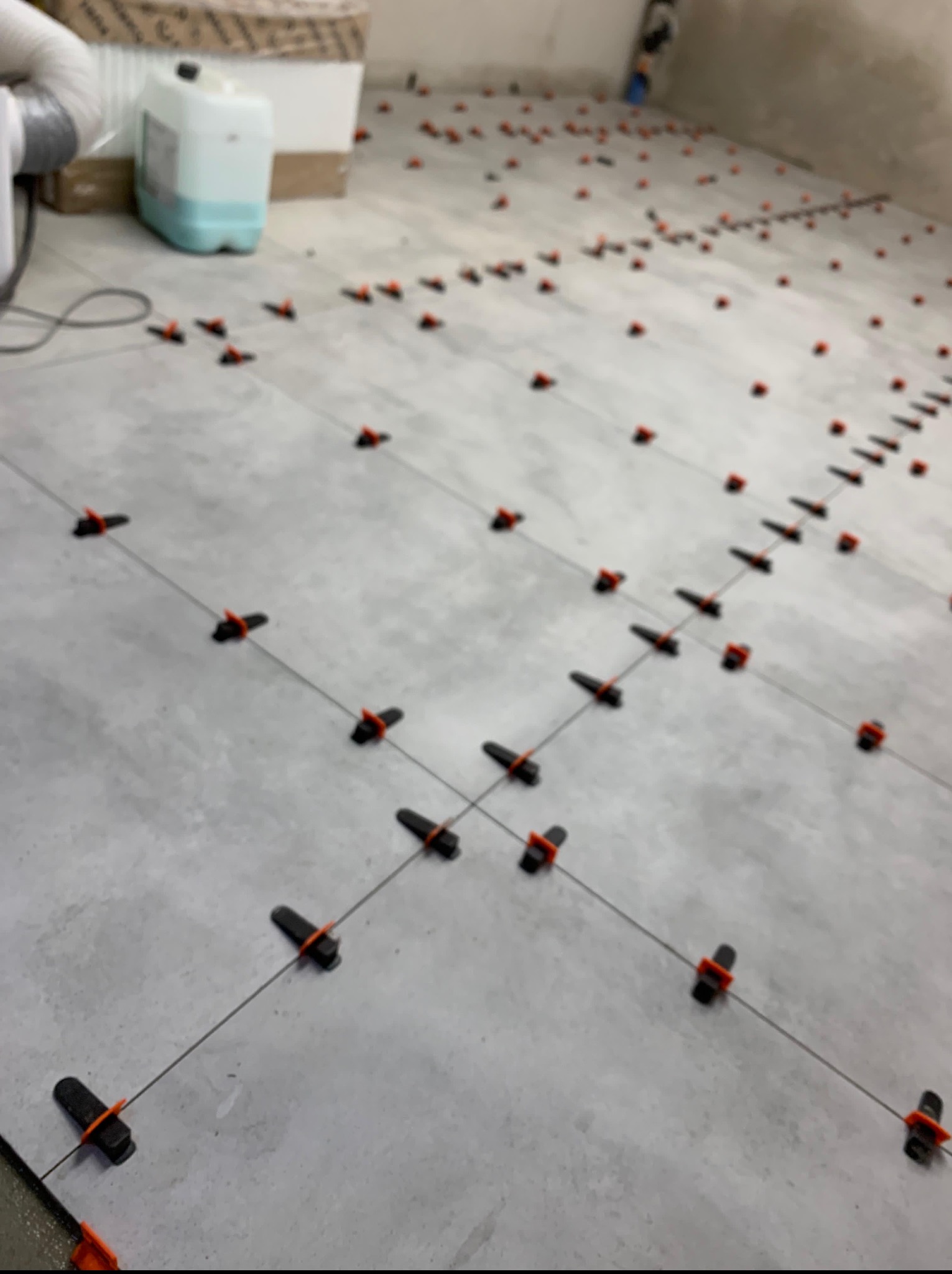 Szare płytki podłogowe, w trakcie układania, z użyciem klipsów i klinów poziomujących. Widoczne narzędzia i materiały budowlane w tle. Wnętrze w trakcie remontu.