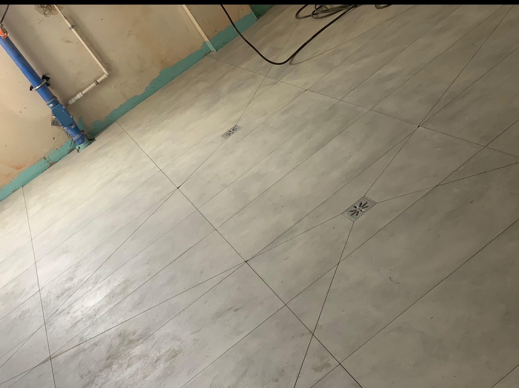 Ukończona podłoga z jasnych, kwadratowych płytek, z widocznymi odpływami liniowymi i fragmentem instalacji wodnej przy ścianie, w trakcie prac wykończeniowych.