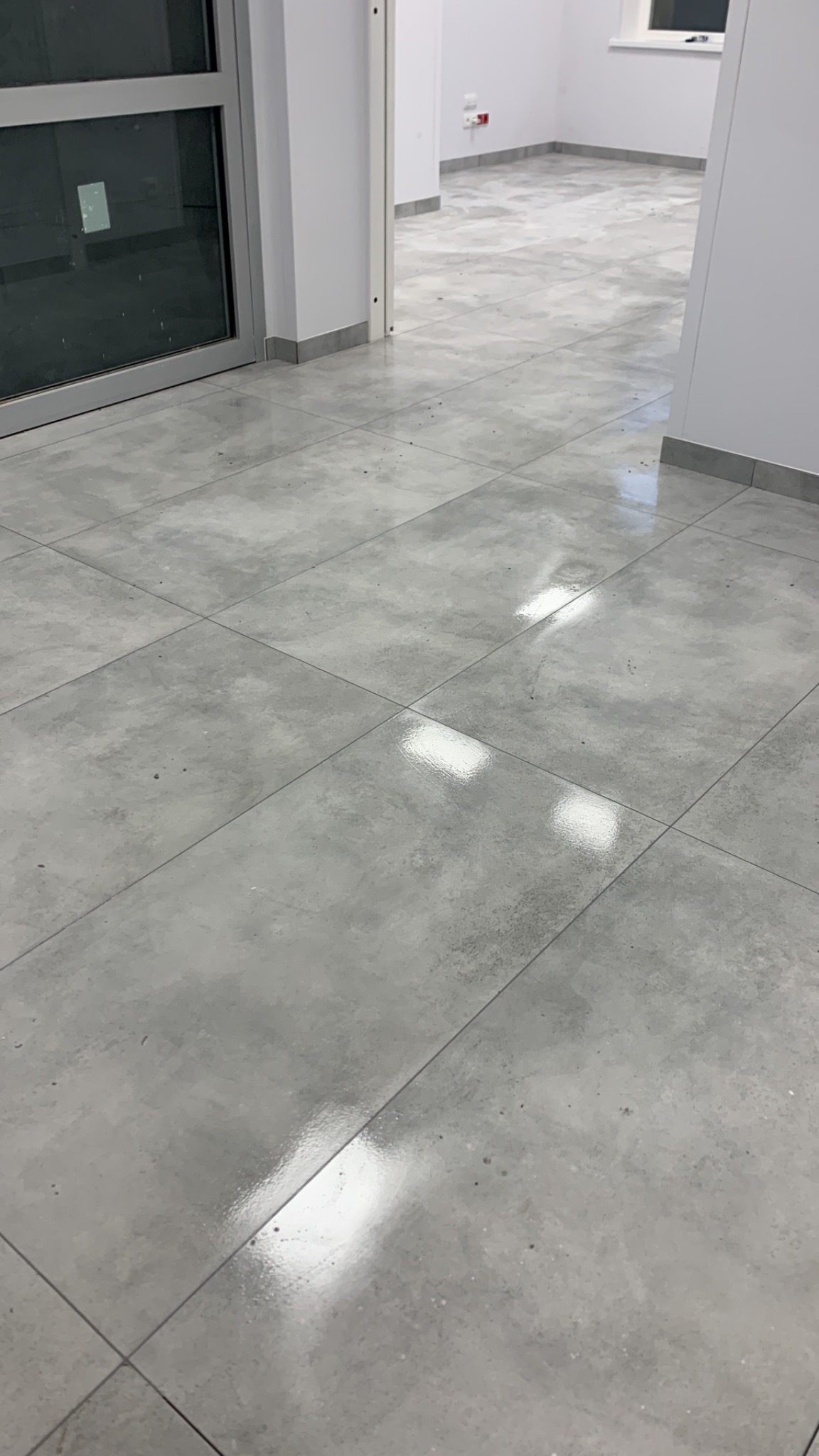 Jasnoszara, błyszcząca podłoga z płytek w nowym pomieszczeniu, widok z niskiej perspektywy, fuga w kolorze zbliżonym do płytek, drzwi z szybą.