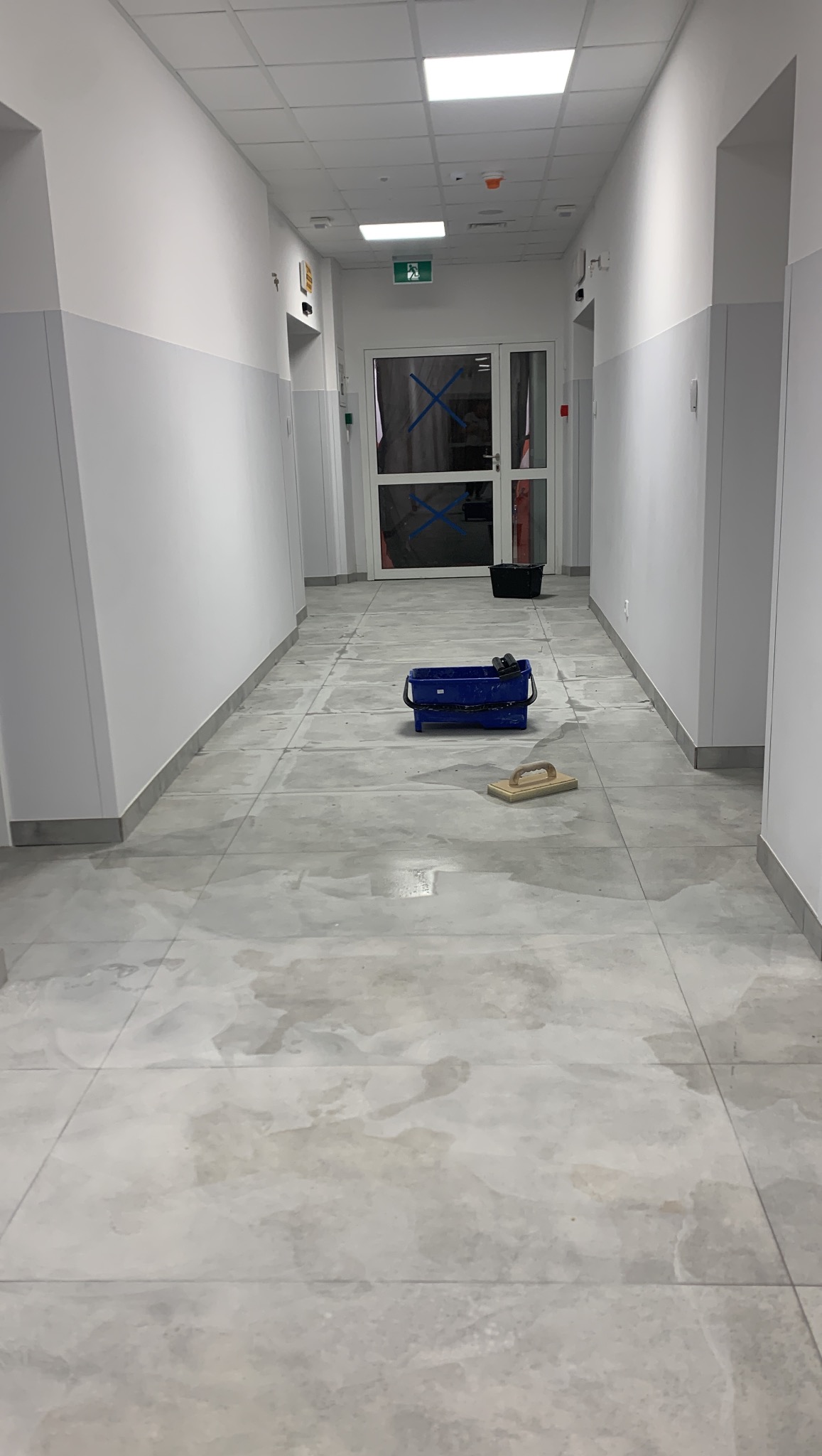 Szary gres szkliwiony ułożony w korytarzu, widoczny sprzęt glazurniczy: wiadro i packa. Świeżo położona podłoga, widoczne ślady wody po myciu fug.