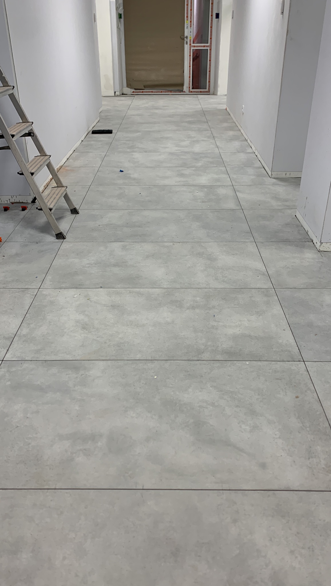 Ułożona szara podłoga z płytek w korytarzu, widoczna drabina malarska i elementy poziomowania płytek. Minimalistyczny styl, jasne wnętrze w trakcie prac wykończeniowych.