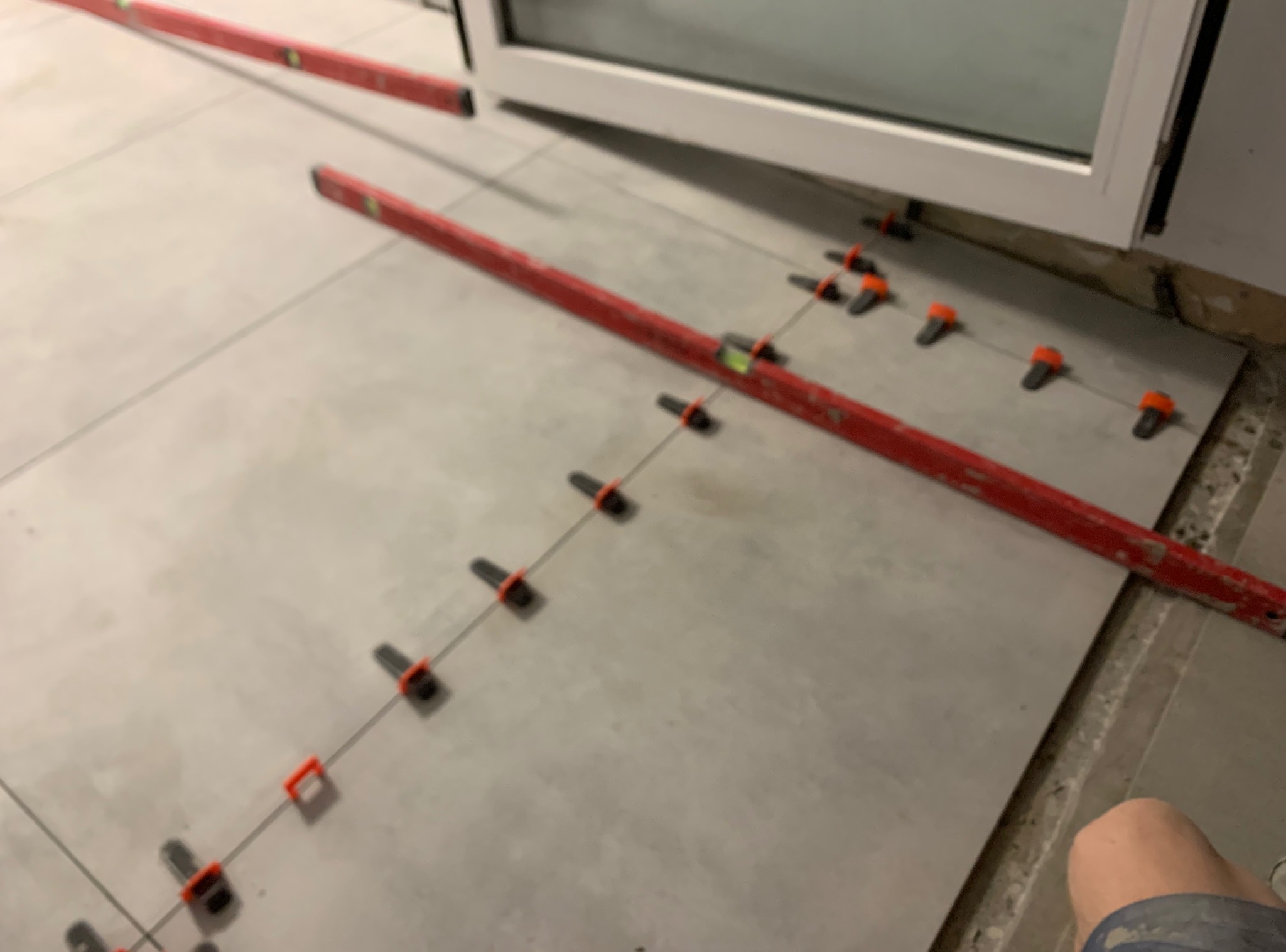 Układanie jasnoszarych płytek podłogowych z użyciem poziomicy i klipsów dystansowych, widok z góry, częściowo widoczne okno balkonowe.