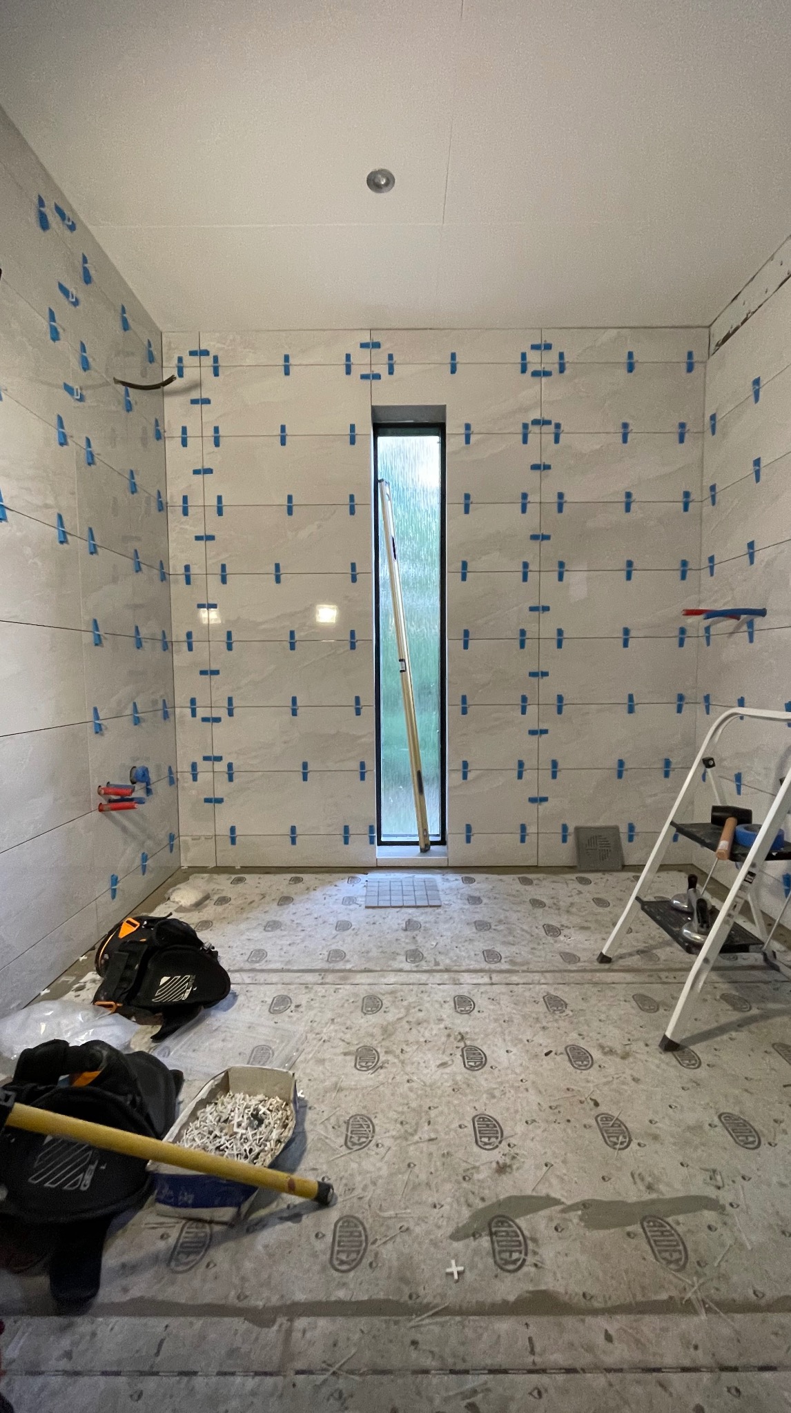 Łazienka w trakcie układania jasnych, poziomych płytek z użyciem niebieskich krzyżyków dystansowych. Widoczne narzędzia i drabina.