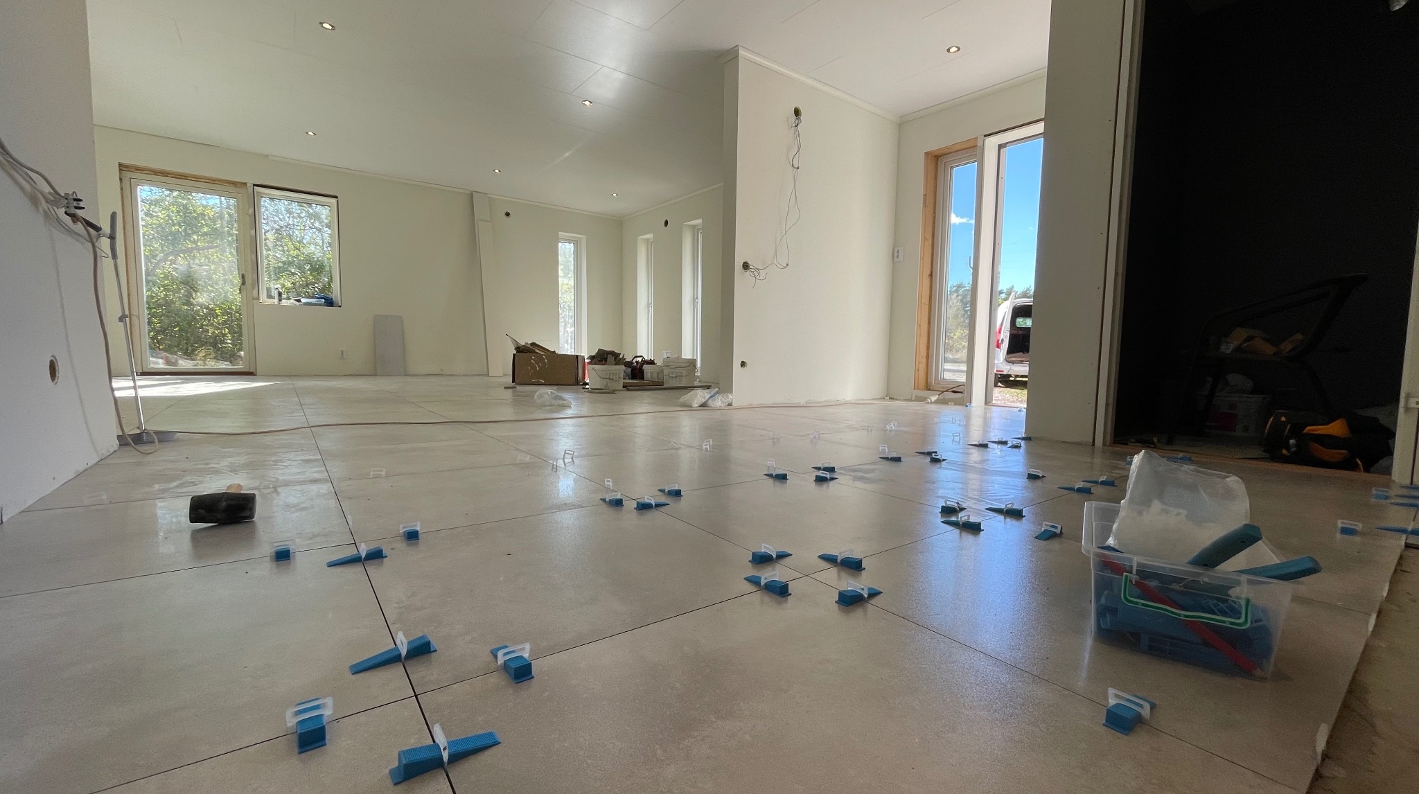 Jasne wnętrze z ułożonymi płytkami podłogowymi, z użyciem systemu poziomowania. Widoczne narzędzia i akcesoria glazurnicze. Duże okna wpuszczają naturalne światło.