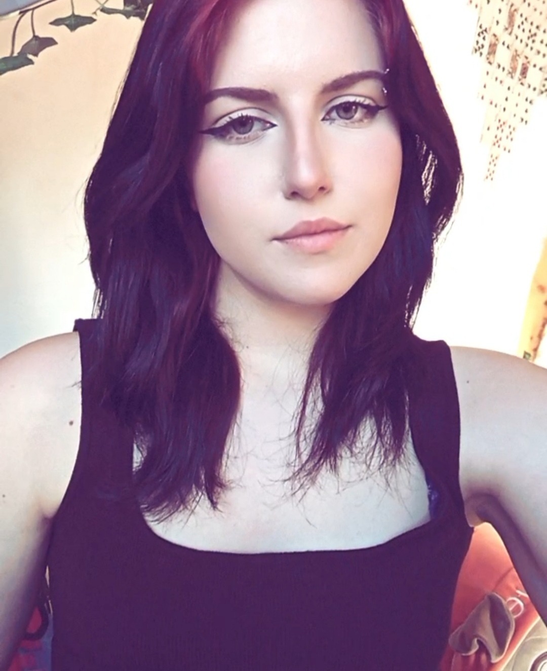 Portret młodej kobiety z ciemnymi włosami i wyrazistym makijażem oczu, ubrana w czarny top na tle jasnej ściany z dekoracjami.