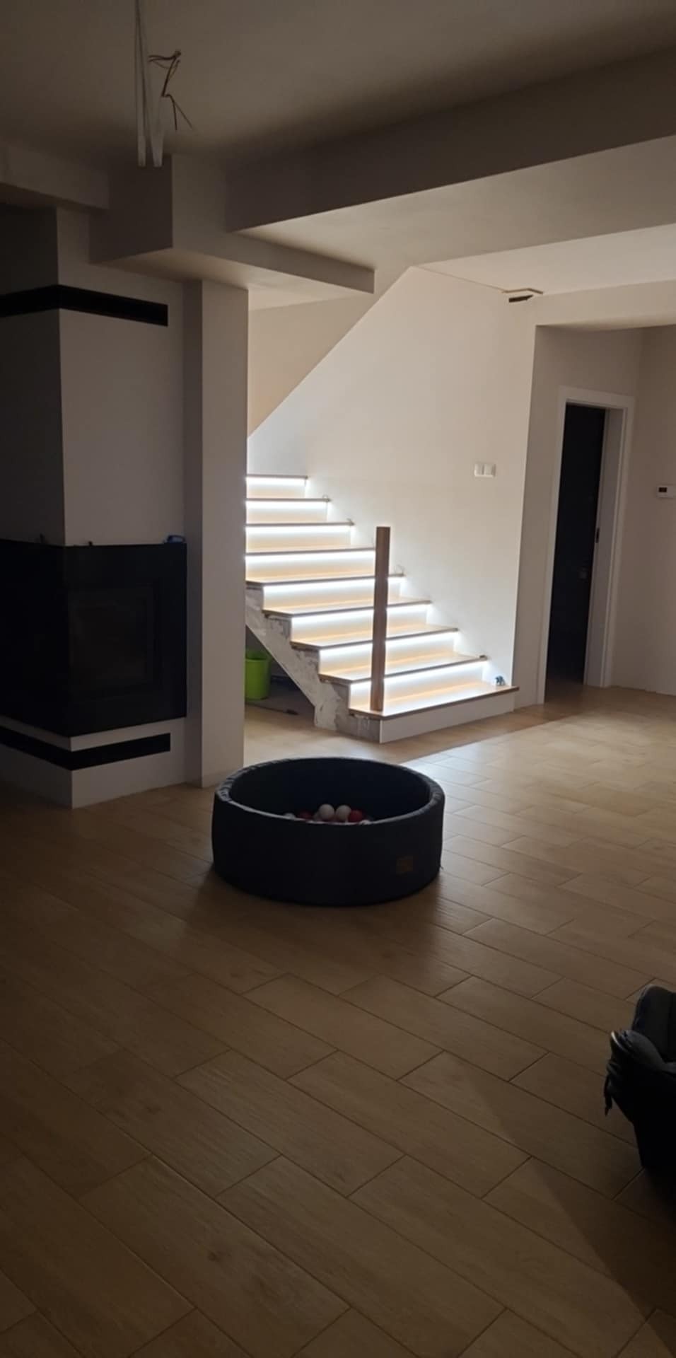 Nowoczesne schody z podświetleniem LED w jasnym wnętrzu domu. Widoczny fragment kominka i podłogi z płytek imitujących drewno. W tle otwarte drzwi.