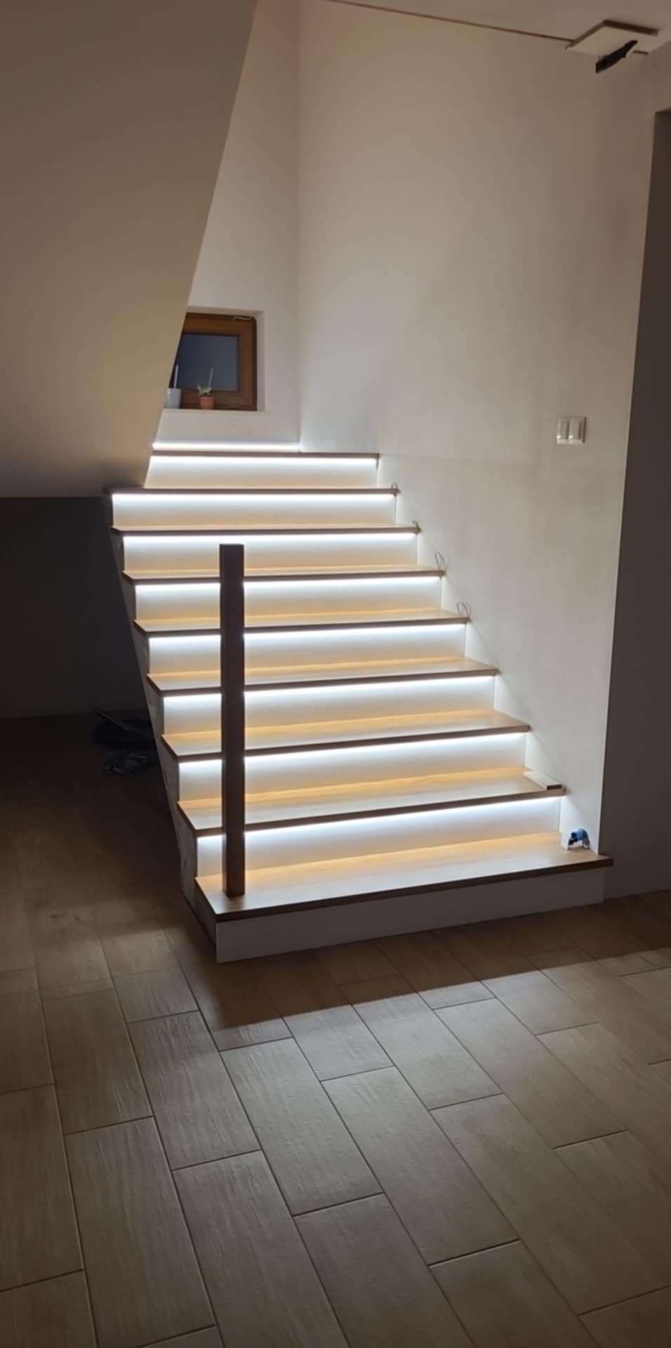 Nowoczesne schody z podświetleniem LED na każdym stopniu, drewniana poręcz. Małe okno z rośliną w tle. Oświetlenie sufitowe. Widok z dołu na jasne wnętrze.