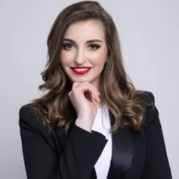 Portret uśmiechniętej kobiety w czarnej marynarce, z dłonią pod brodą, na jasnym tle. Wyraziste usta pomalowane czerwoną szminką. Profesjonalny wizerunek.