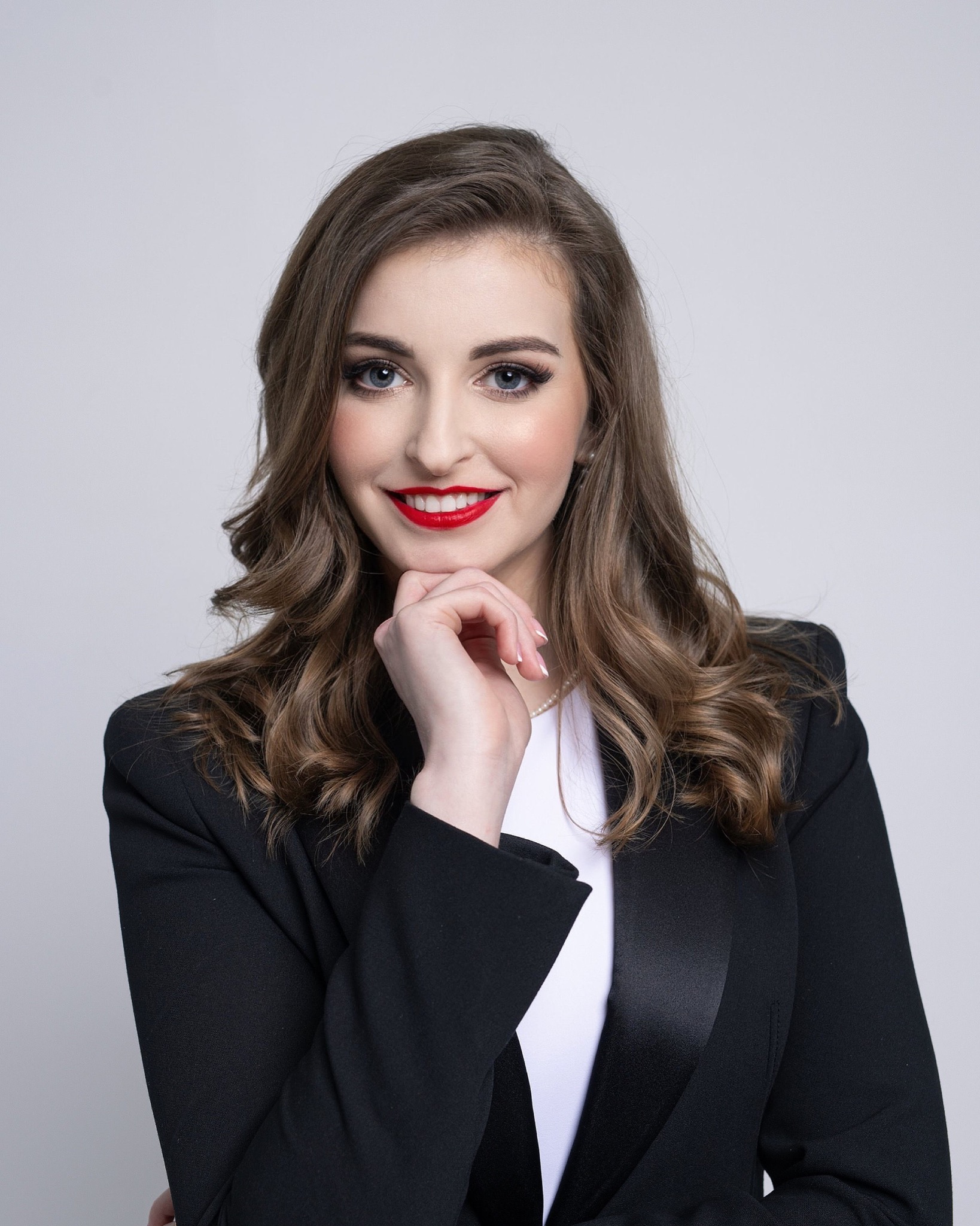 Portret uśmiechniętej kobiety w czarnej marynarce, z dłonią pod brodą, na jasnym tle. Wyraziste usta pomalowane czerwoną szminką. Profesjonalny wizerunek.