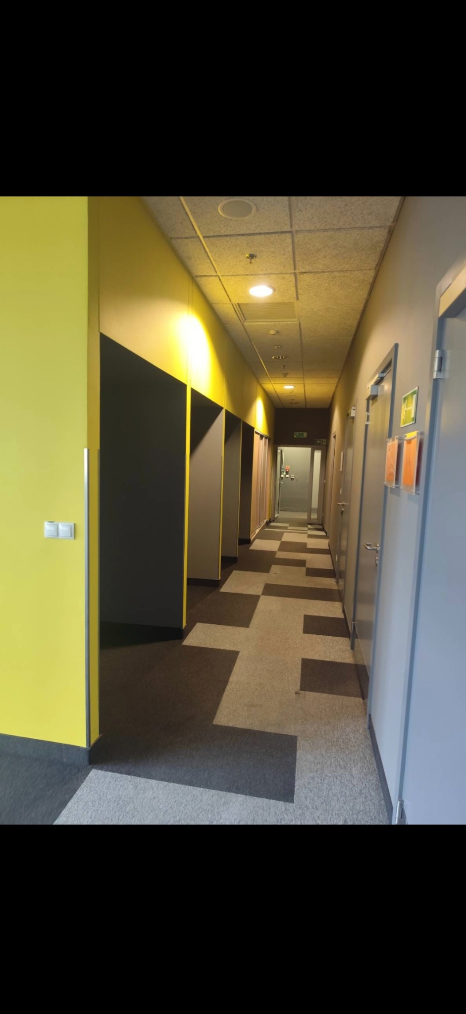 Długi korytarz biurowy z szaro-żółtą kolorystyką ścian i podłogą w szachownicę. Widoczne drzwi i oświetlenie sufitowe.