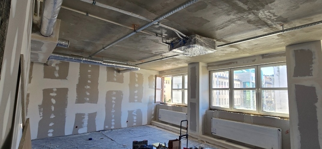 Wnętrze w trakcie prac wykończeniowych: ściany pokryte gładzią, widoczne instalacje pod sufitem, okna z widokiem na miasto. Przygotowanie do dalszych prac.