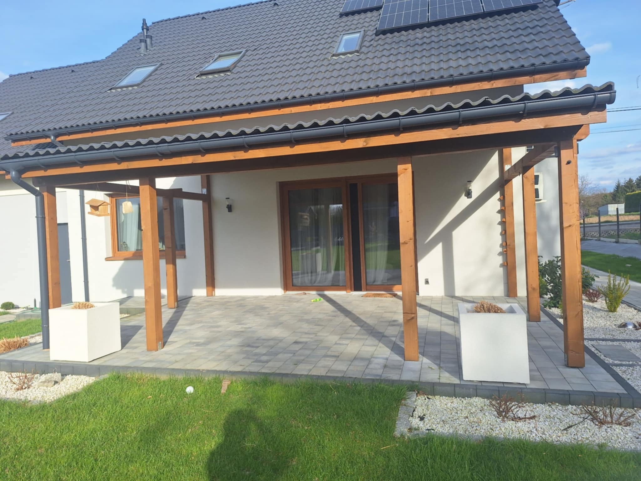Nowoczesny dom z drewnianą pergolą i szarą kostką brukową na tarasie. Na dachu panele słoneczne, a w tle błękitne niebo. Widok z poziomu trawnika.