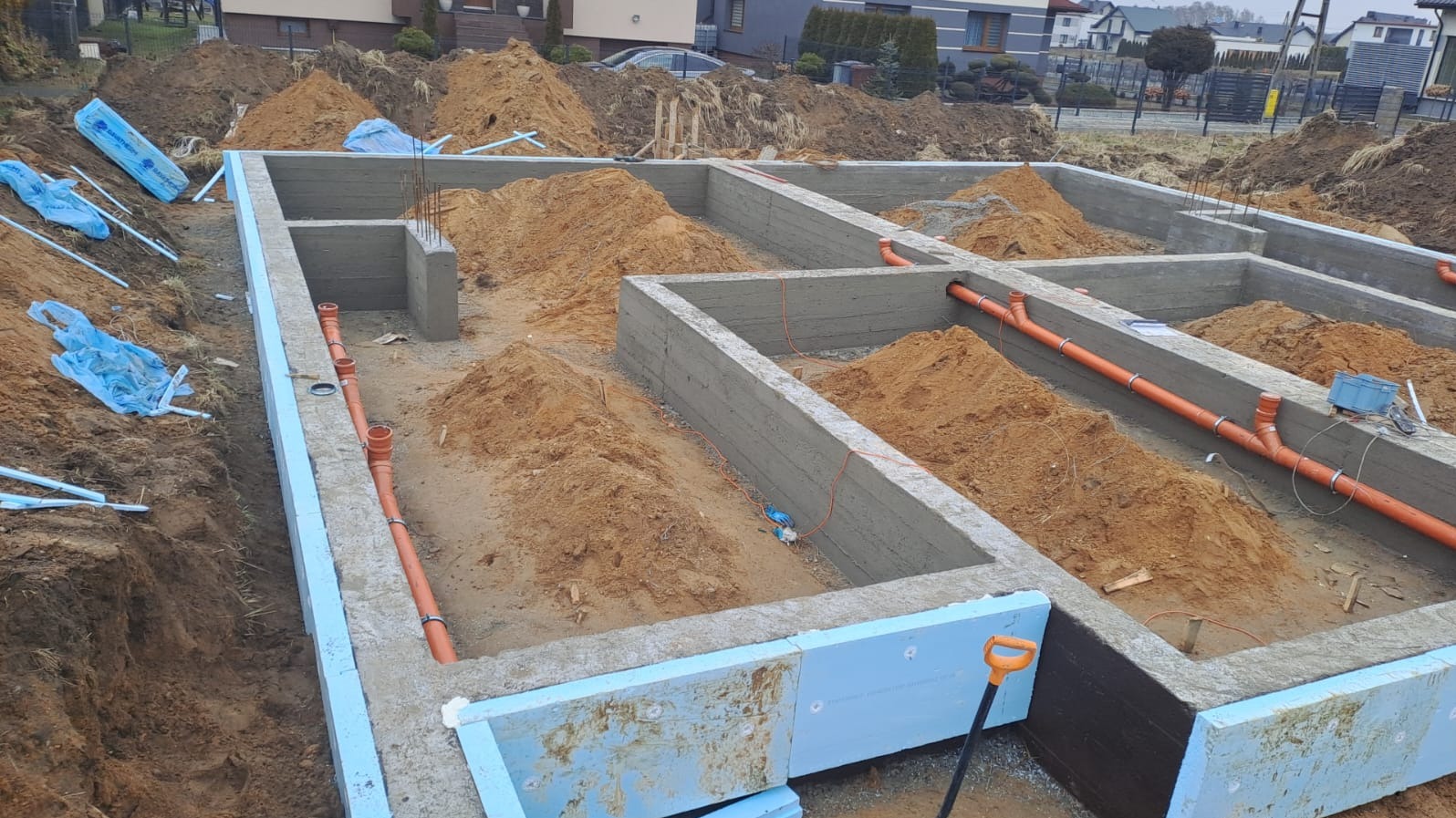 Zalane fundamenty domu z izolacją termiczną i instalacją kanalizacyjną w wykopie, przygotowane do dalszych prac budowlanych. Widoczne elementy zbrojenia i rury kanalizacyjne.