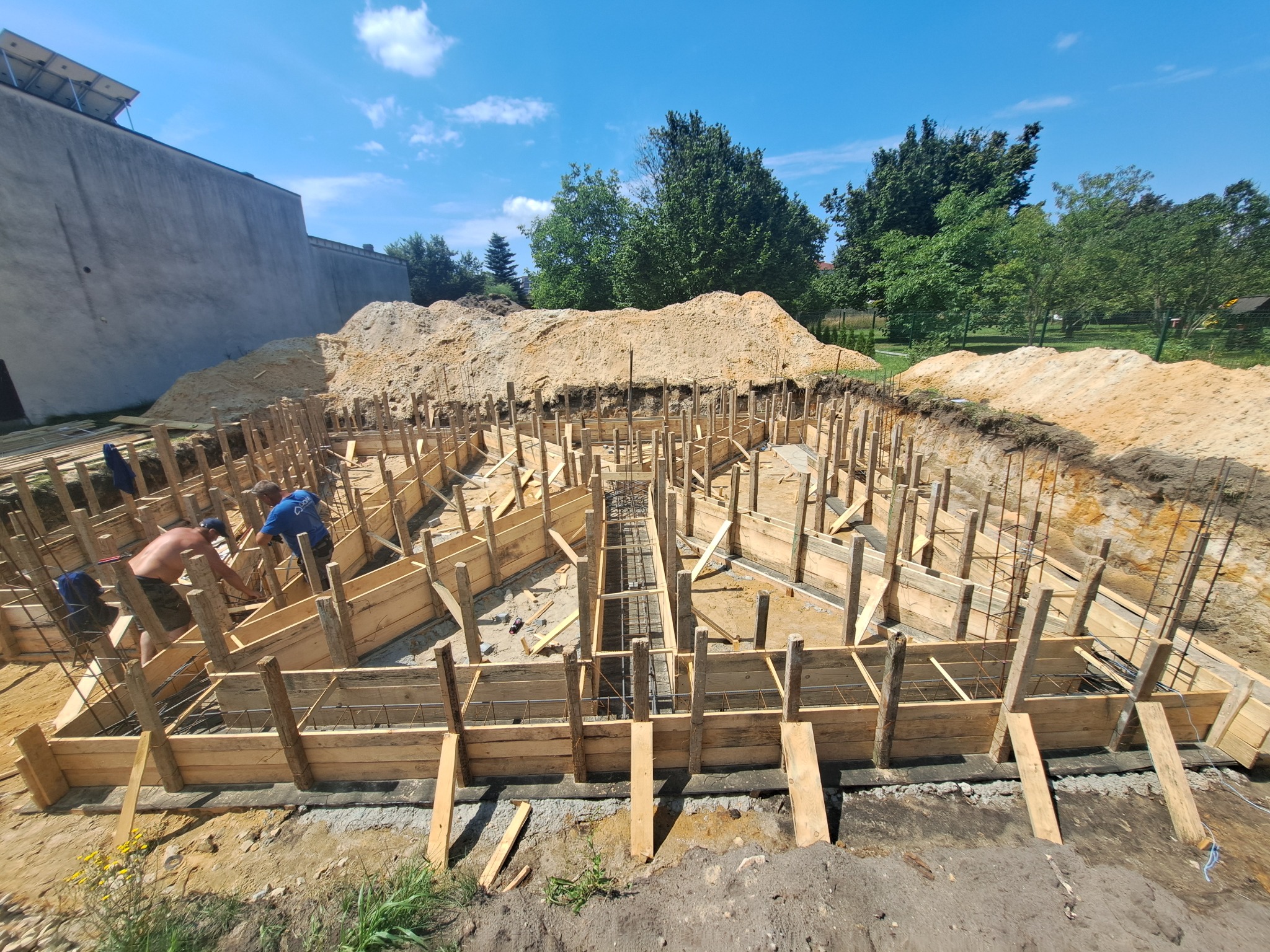 Szeroki kadr na drewniane szalunki fundamentów zbrojonych stalą, z pracownikami w tle, pod błękitnym niebem i stertą piasku. Widok na plac budowy.