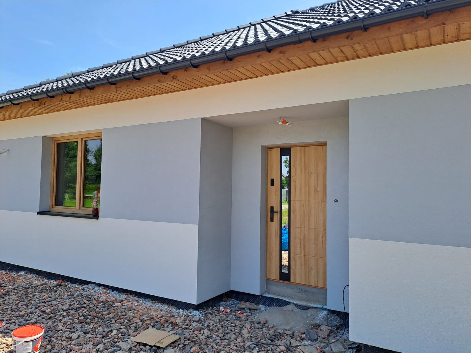 Elewacja domu jednorodzinnego w odcieniach szarości i bieli, z drewnianą stolarką okienną i nowoczesnymi drzwiami wejściowymi. Widoczny fragment dachu i otoczenie budynku.