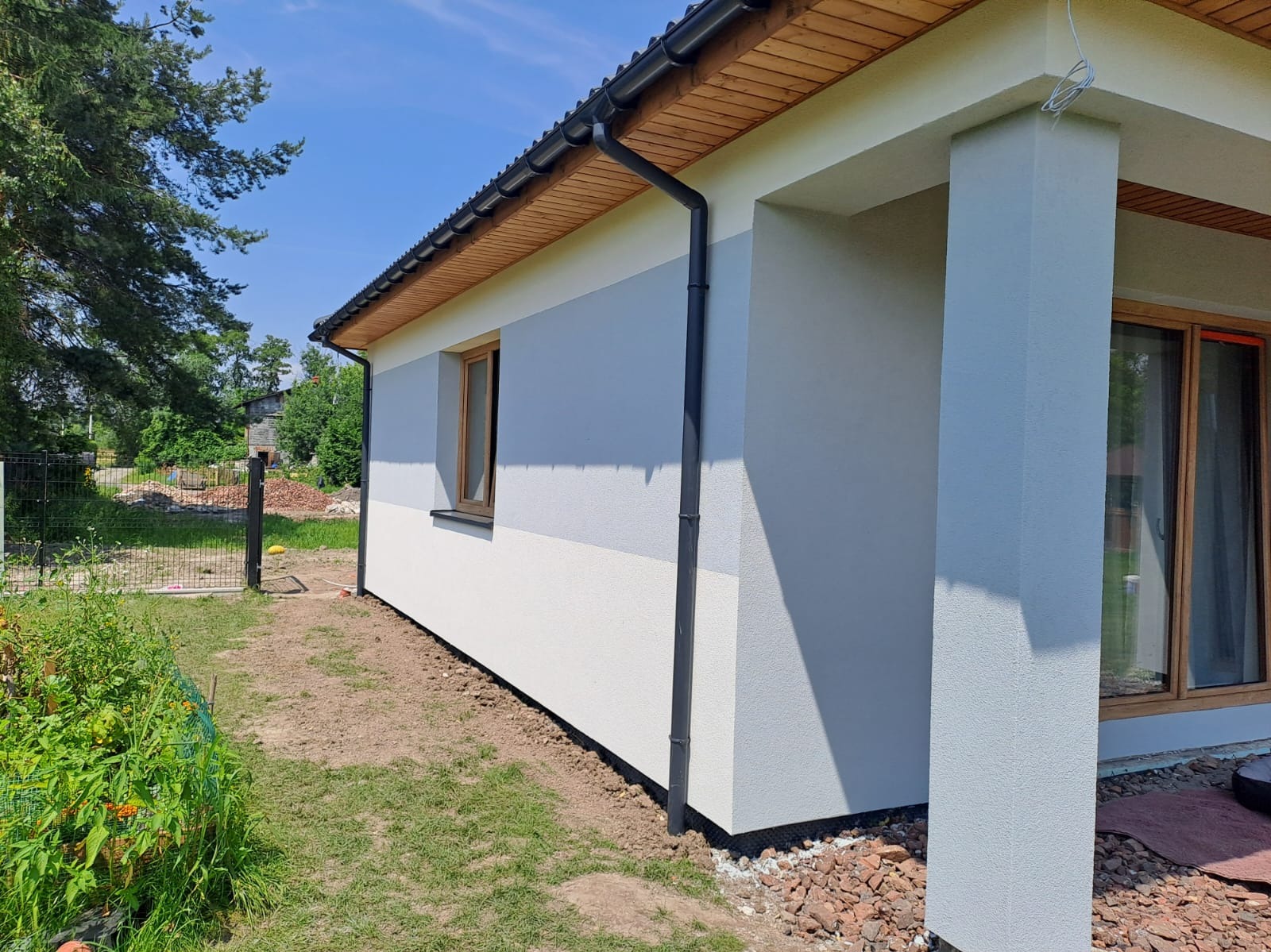 Wykończony dom jednorodzinny z dwukolorową elewacją: szarym pasem na górze i białym na dole. Widoczny fragment dachu z drewnianą podbitką i rynną. Dom z oknami w drewnianych ramach.
