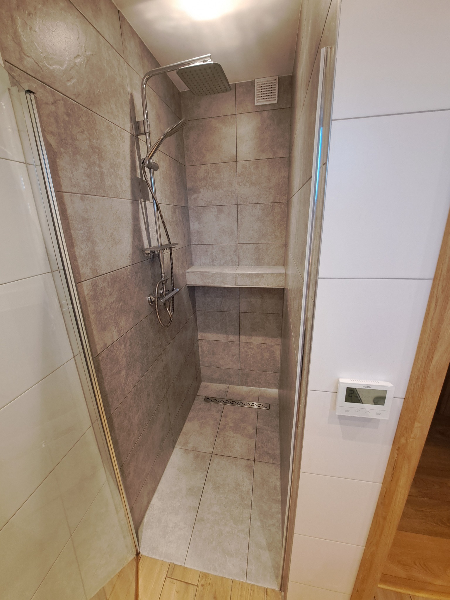 Nowoczesna kabina prysznicowa z brązowymi płytkami imitującymi kamień, półką i odpływem liniowym. Białe ściany i sterownik temperatury dopełniają wystrój.