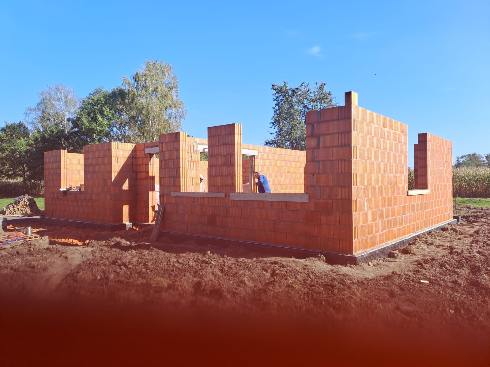 Wznoszenie ścian domu z czerwonej cegły na fundamencie. Widoczny pracownik w niebieskiej koszulce. W tle drzewa i błękitne niebo.
