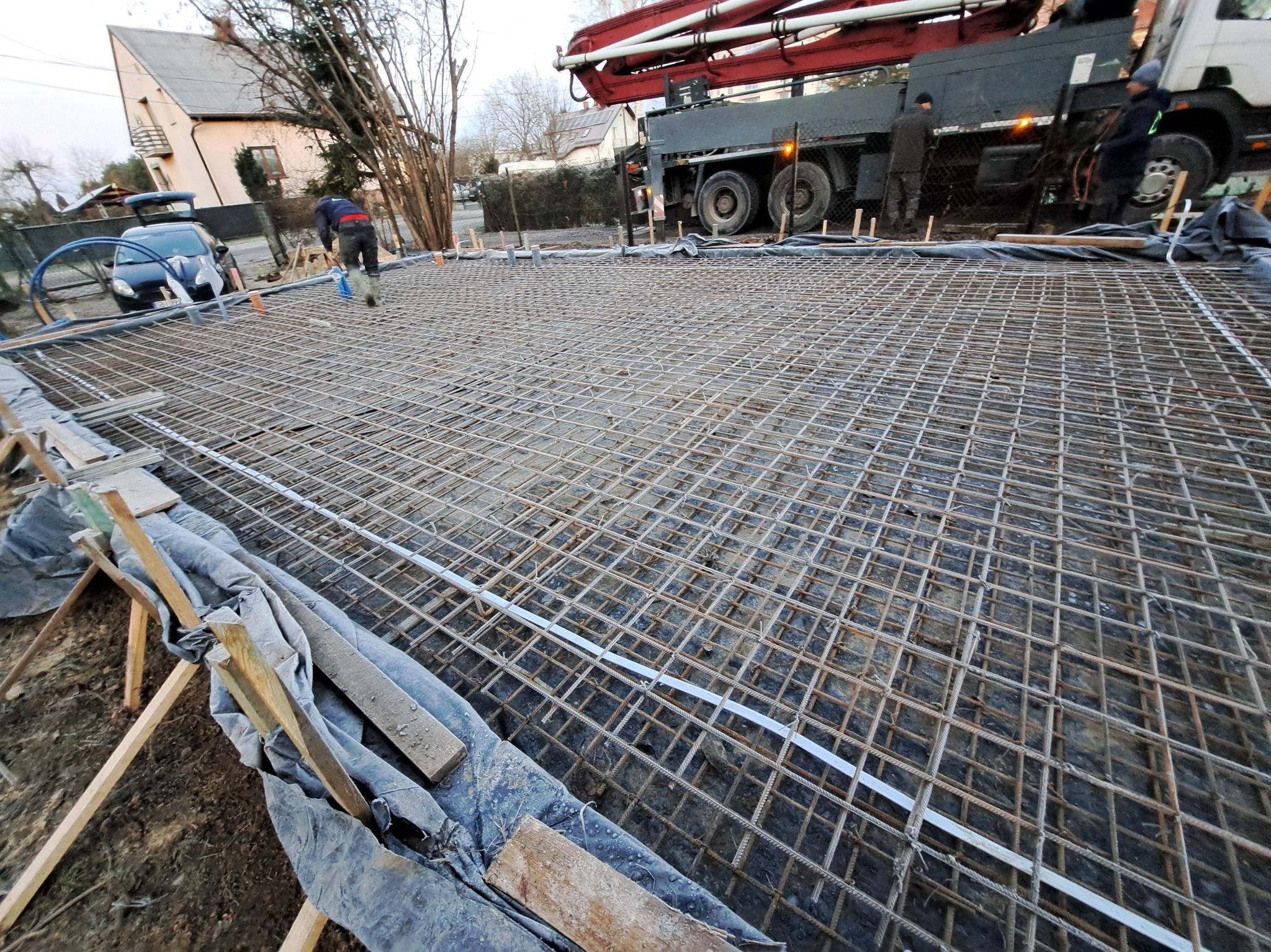 Zbrojenie fundamentu domu jednorodzinnego, widok z góry na siatkę stalową, w tle betonowóz i pracownicy. Przygotowanie do wylewania betonu.