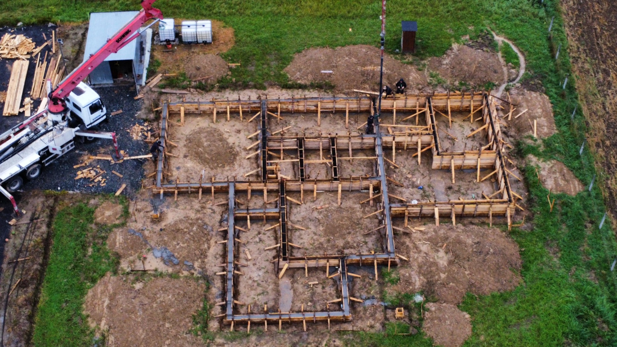 Z góry: szalunek fundamentów domu jednorodzinnego na działce, z widocznym dźwigiem i pracownikami. W tle zielona trawa i drzewa. Etap przygotowania do wylania betonu.