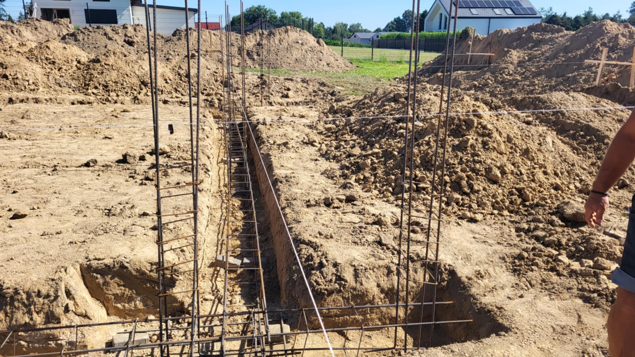 Zbrojenie wykopu pod fundamenty domu jednorodzinnego, widoczne stosy ziemi i fragment budynku w tle. Kąt ujęcia z dołu podkreśla głębokość wykopu.