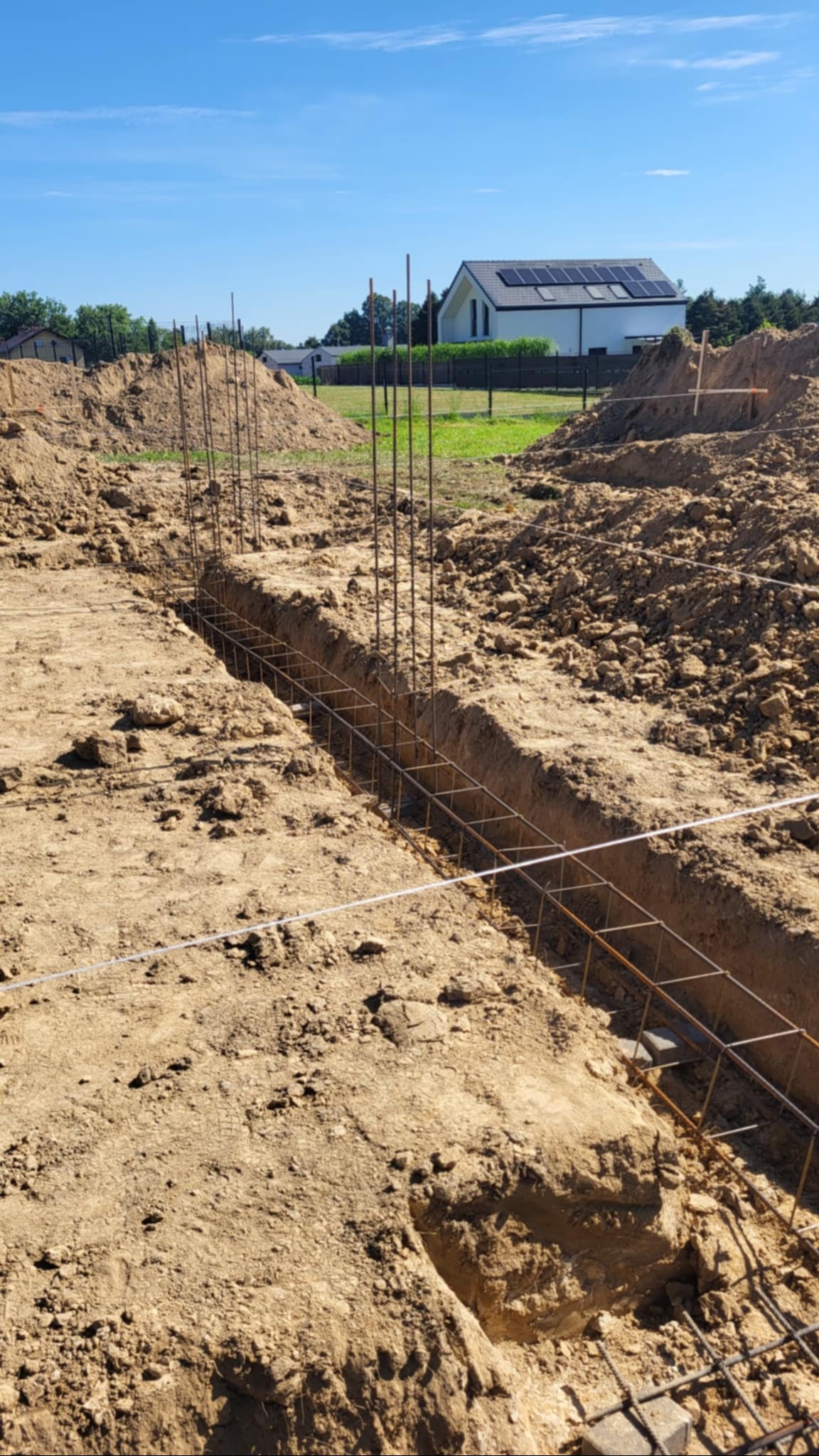 Zbrojenie fundamentów w wykopie na tle domu z panelami słonecznymi. Widoczne pionowe pręty i poziome siatki z drutu. Ziemia usypana wokół wykopu.