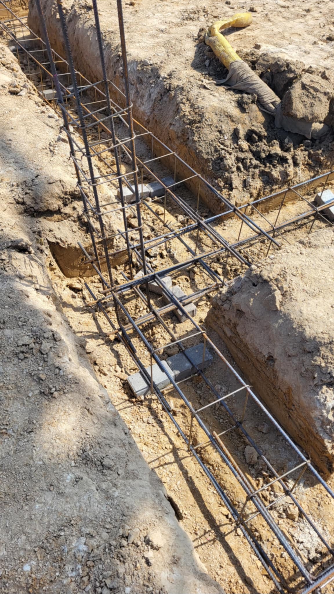 Zbrojenie fundamentów w wykopie, z użyciem stalowych prętów i betonowych dystansów. Widoczny fragment żółtego węża. Jasne oświetlenie podkreśla surowość materiałów.