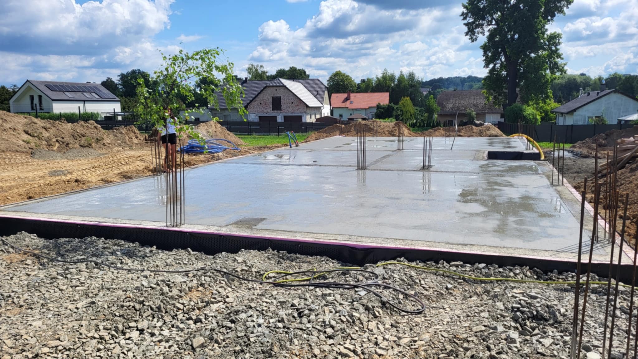 Wylana płyta fundamentowa z wystającymi prętami zbrojeniowymi na działce budowlanej w słoneczny dzień. Widoczne izolacje i przygotowany teren pod budowę.