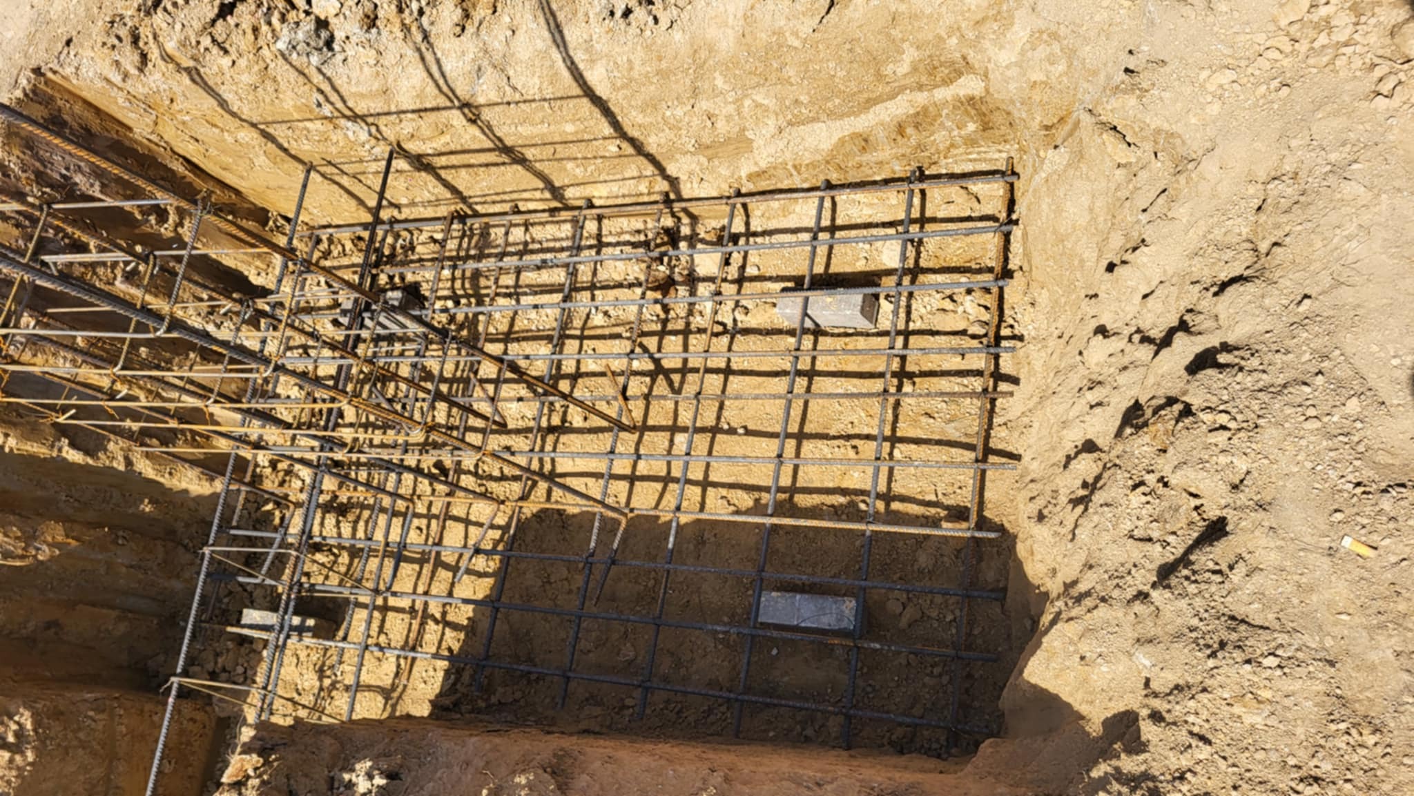 Zbrojenie fundamentu w wykopie, widok z góry. Konstrukcja z prętów stalowych tworzy siatkę. Fragmenty cegieł użyte jako podkładki. Cienie rzucane przez słońce.
