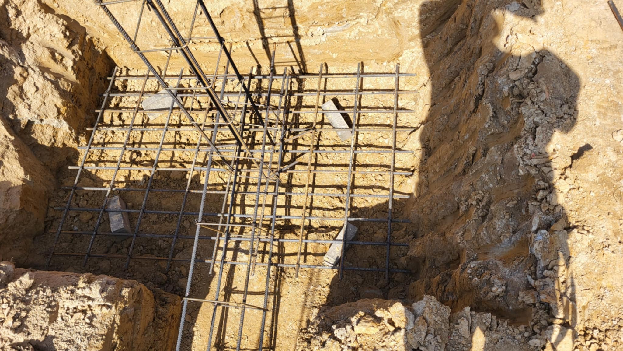 Zbrojenie fundamentu w wykopie, widoczne stalowe pręty tworzące siatkę, podparte kamieniami. Ziemia w wykopie oświetlona słońcem, cień osoby w prawym górnym rogu.
