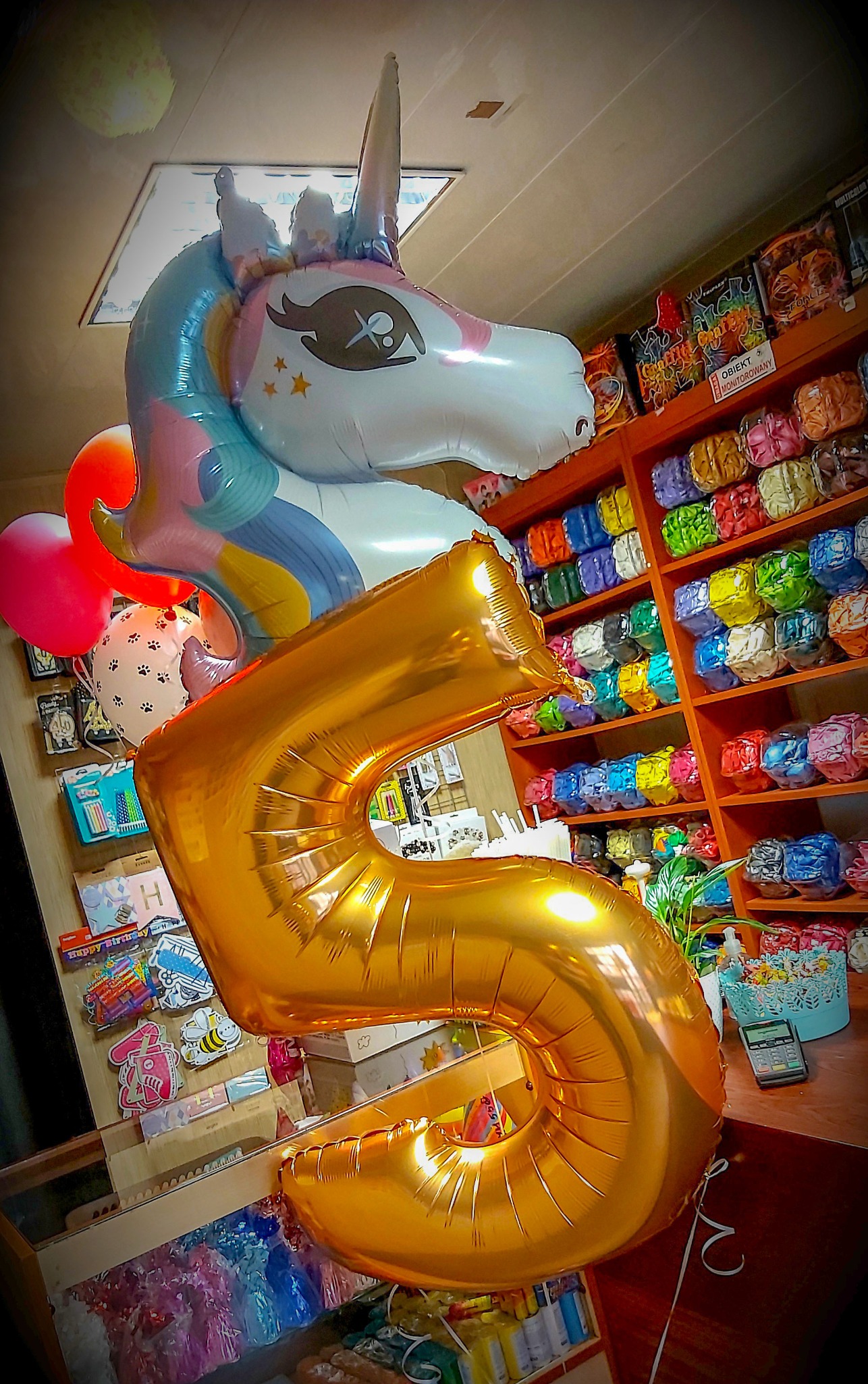 Zestaw balonów: złota cyfra 5, jednorożec z tęczową grzywą i balony z motywem łapek, na tle regału z kolorowymi balonami i akcesoriami imprezowymi.