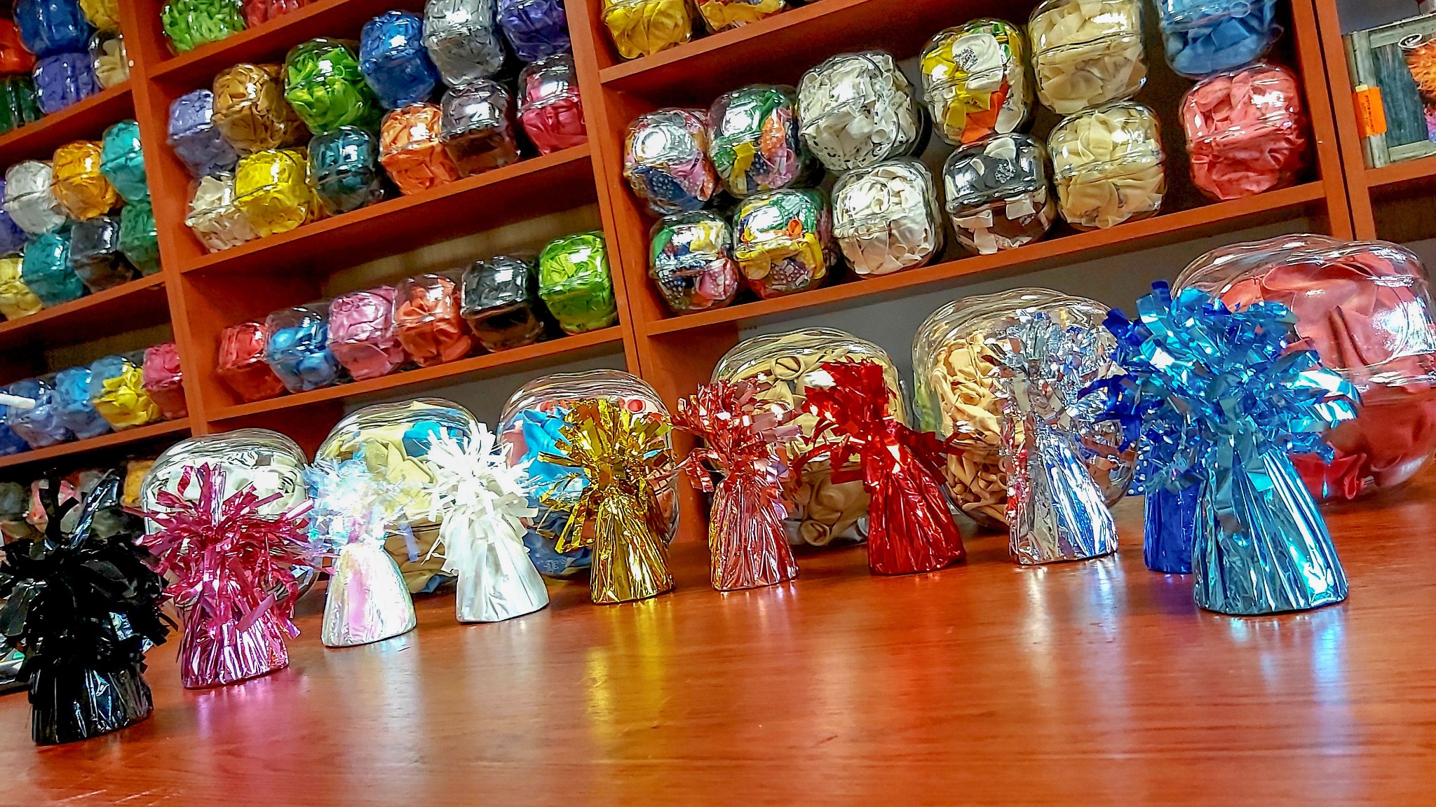 Kolorowe balony zapakowane w przezroczyste słoiki i obciążniki z błyszczącej folii, ustawione na drewnianej półce i blacie.