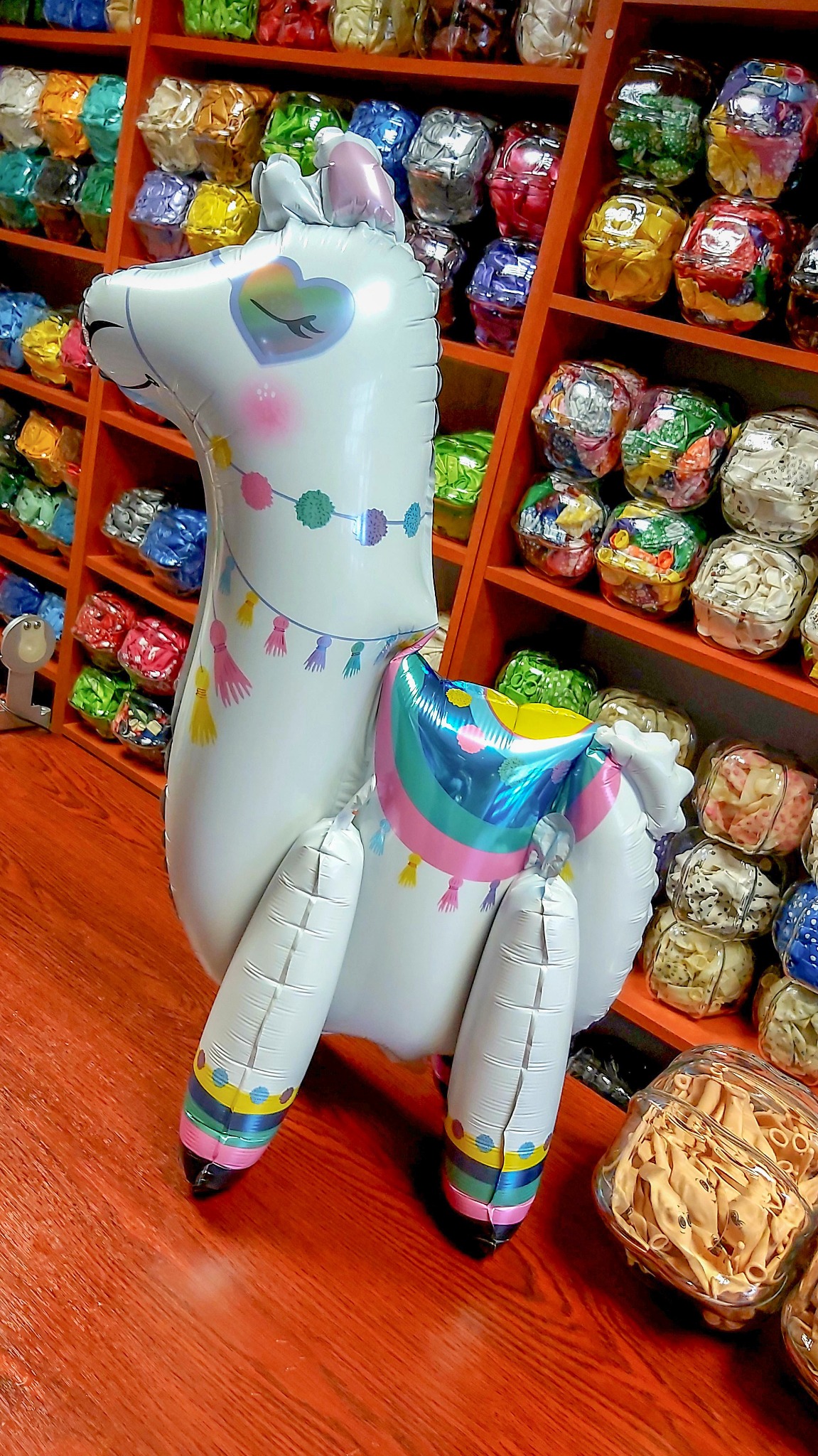 Duży, biały balon w kształcie lamy z tęczowym sercem i kolorowymi frędzlami, stojący na pomarańczowej podłodze, na tle półek wypełnionych opakowaniami z balonami.