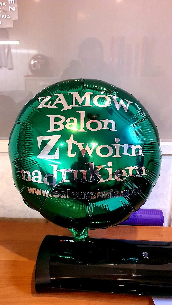 Błyszczący, zielony balon foliowy z srebrnym nadrukiem 'Zamów Balon Z Twoim nadrukiem' i adresem strony internetowej, ustawiony na biurku.