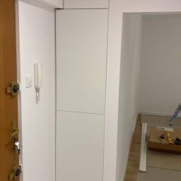 Daniel Dzierbicki - Wysoka, biała szafa wnękowa z gładkimi frontami, zamontowana przy wejściu, obok drewnianych drzwi. Widoczne marmurowe płytki podłogowe i fragment wnętrza pokoju.