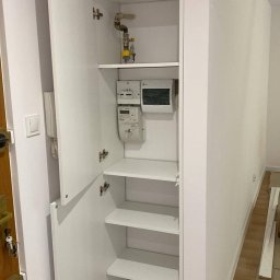 Daniel Dzierbicki - Biała szafa wnękowa z otwartymi drzwiami, widoczne półki, instalacja gazowa i liczniki. Marmurowa podłoga, drzwi wejściowe po lewej stronie.