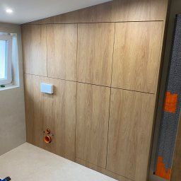 Daniel Dzierbicki - Drewniana zabudowa ściany w łazience z ukrytą spłuczką i instalacją sanitarną. Widoczne płytki podłogowe i fragment okna. Jasne oświetlenie.