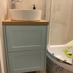 Daniel Dzierbicki - Błękitna szafka łazienkowa z drewnianym blatem, na którym stoi biała umywalka nablatowa i dozownik mydła. W tle kabina prysznicowa z widocznym siedziskiem dla dziecka.