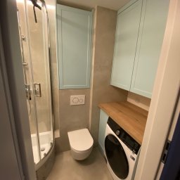 Daniel Dzierbicki - Funkcjonalna łazienka z kabiną prysznicową, toaletą i pralką zabudowaną pod drewnianym blatem, nad którą wiszą szafki w kolorze pastelowego błękitu.