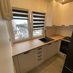 Daniel Dzierbicki - Jasna kuchnia z drewnianym blatem i czarnymi akcentami: bateria, zlew, uchwyty. Za oknem widok na miasto w zimowej scenerii. Nowoczesny design.