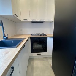 Daniel Dzierbicki - Nowoczesna kuchnia z białymi szafkami, drewnianym blatem i czarnymi akcentami: zlew, bateria, uchwyty. Minimalistyczny design, płyta gazowa i piekarnik w zabudowie.