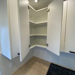 Daniel Dzierbicki - Biała, narożna szafka kuchenna z otwartymi drzwiczkami, ukazująca puste półki. Widoczna płyta gazowa i fragment blatu. Oświetlenie sufitowe.