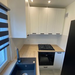 Daniel Dzierbicki - Jasna kuchnia z drewnianym blatem, czarnym zlewem i piekarnikiem. Białe szafki wiszące z minimalistycznymi uchwytami. Widok na okno z roletami dzień-noc.