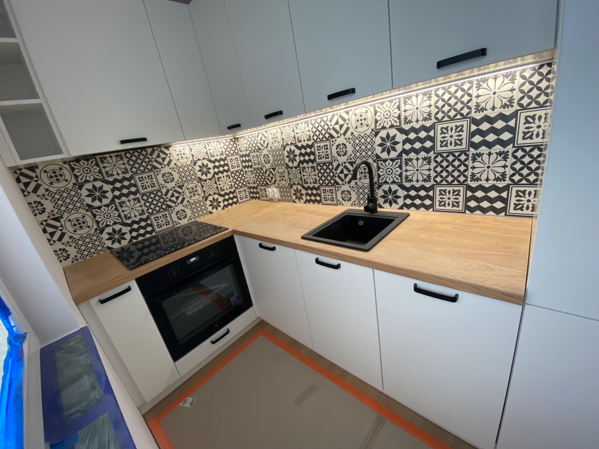 Nowoczesna, biała kuchnia z drewnianym blatem i czarnymi uchwytami. Ściana nad blatem wyłożona czarno-białymi, wzorzystymi płytkami. Widoczny fragment zabezpieczonej podłogi.