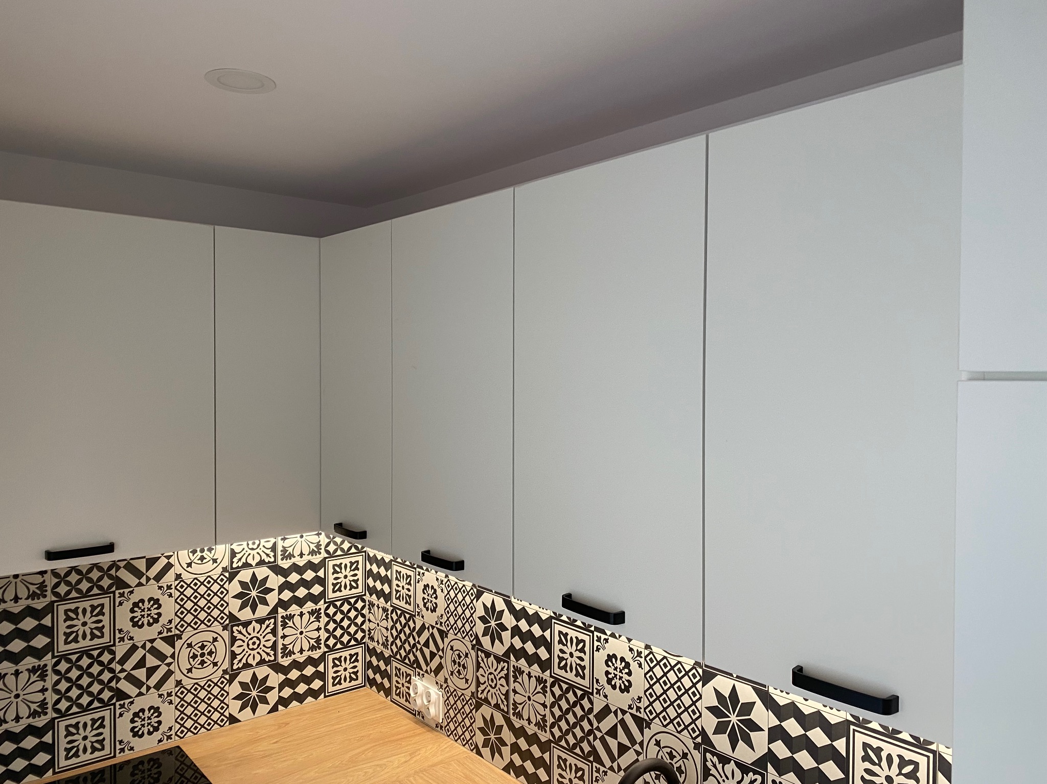 Białe szafki kuchenne z czarnymi uchwytami i wzorzystą mozaiką nad blatem z drewna. Nowoczesny design z elementami retro. Oświetlenie podszafkowe.