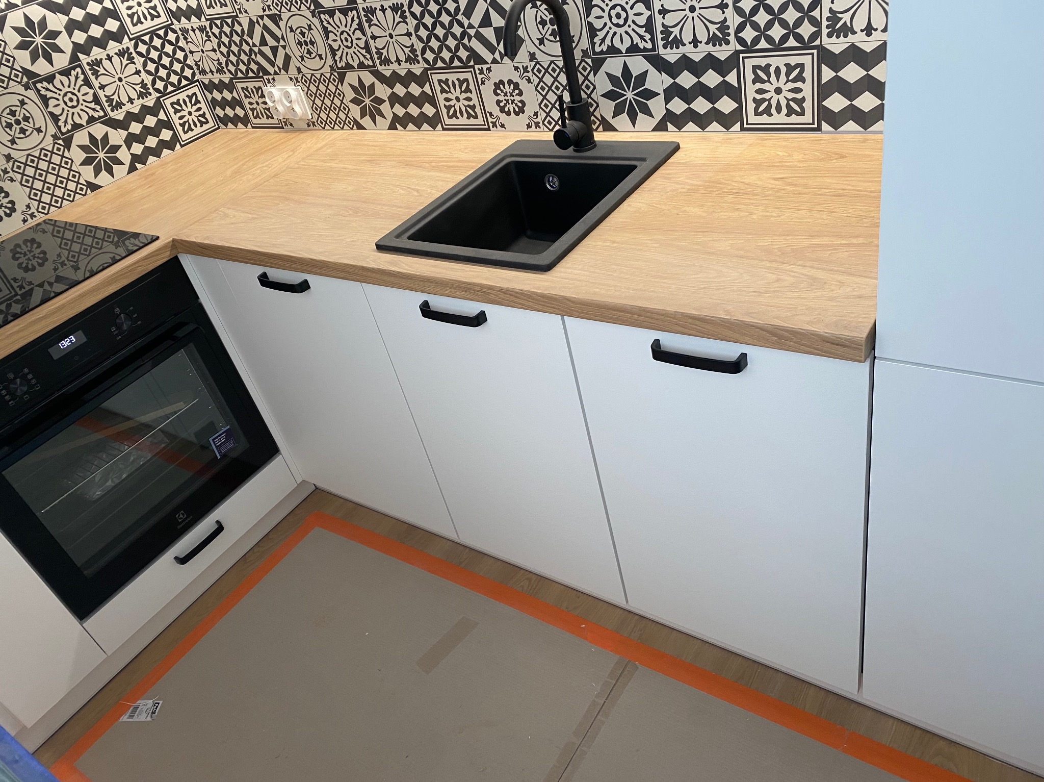 Nowoczesna kuchnia: białe szafki z czarnymi uchwytami, drewniany blat, czarny zlew i bateria. W tle wzorzyste, czarno-białe płytki. Ochrona podłogi.