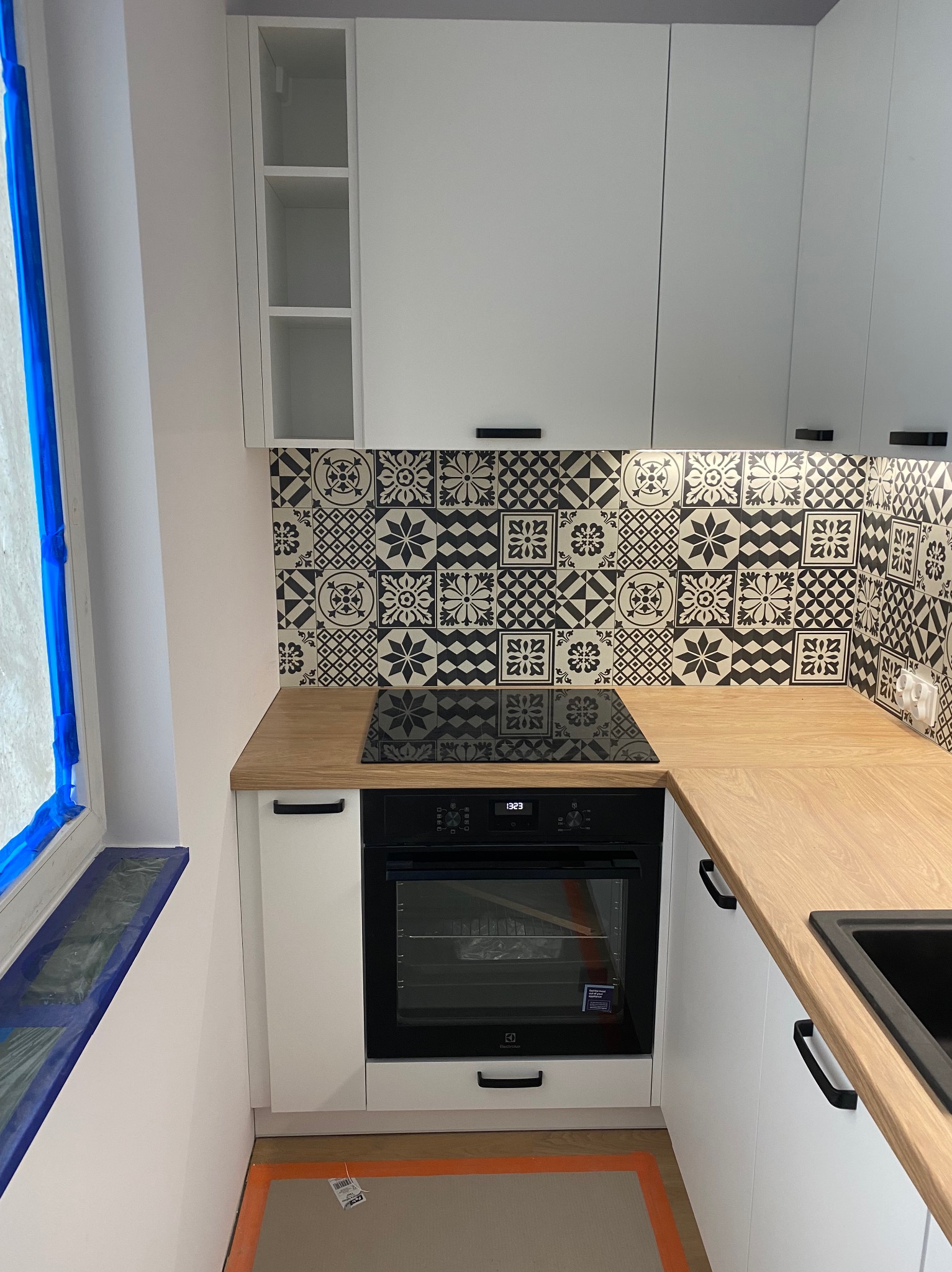 Biała zabudowa kuchenna z drewnianym blatem, czarnymi uchwytami i wzorzystymi płytkami nad blatem. Okno zabezpieczone folią malarską. Nowoczesny design.