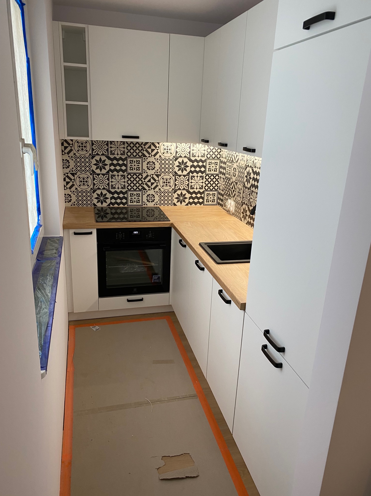 Biała, nowoczesna kuchnia z czarnymi uchwytami i wzorzystą mozaiką nad blatem. Widoczne okno zabezpieczone folią malarską i płyta indukcyjna.