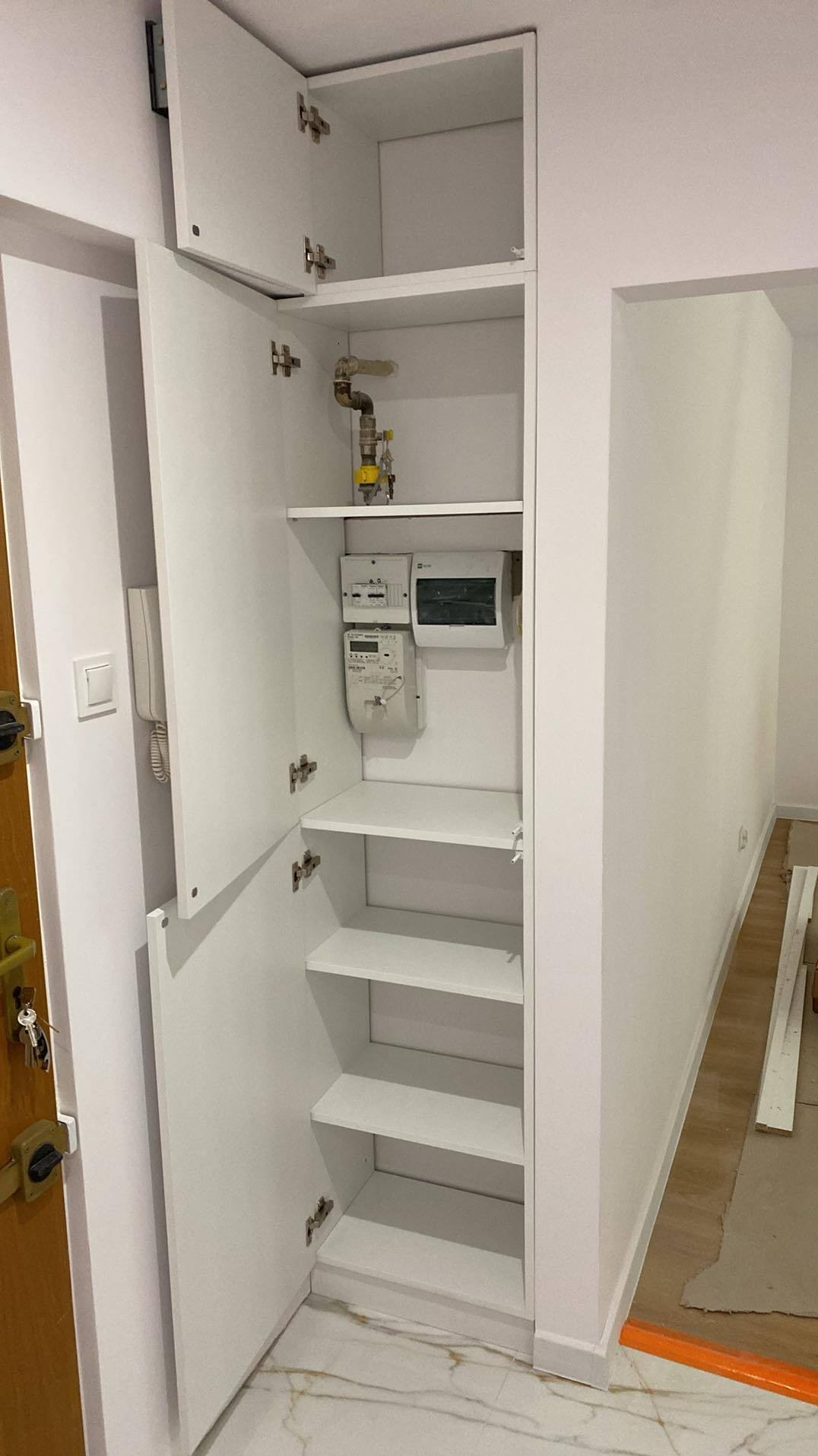 Biała szafa wnękowa z otwartymi drzwiami, widoczne półki, instalacja gazowa i liczniki. Marmurowa podłoga, drzwi wejściowe po lewej stronie.